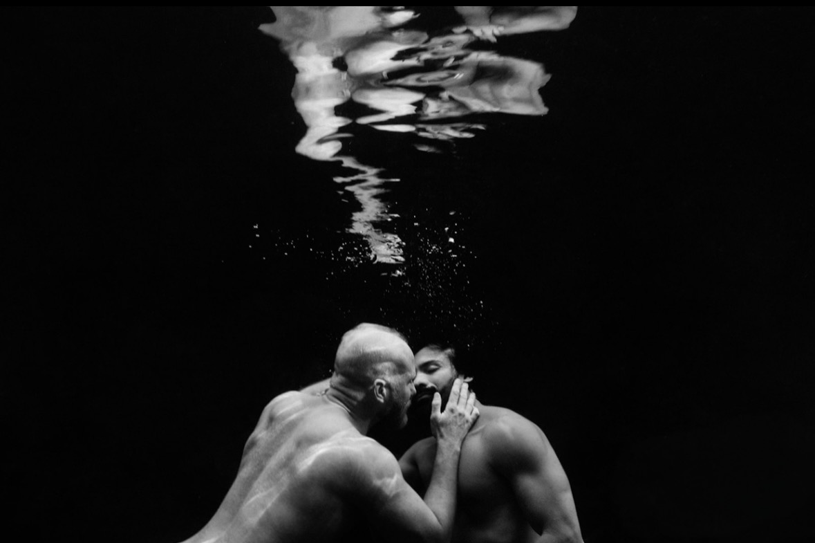 Two men kissing underwater in black and white, light rippling on the surface above
